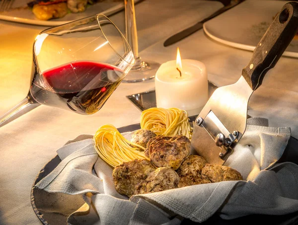 Alba white truffles in napkin with raw egg tagliolini and steel truffle cutter on the restaurant table with burning candle and goblet of red wine, nebbiolo or barbera wine