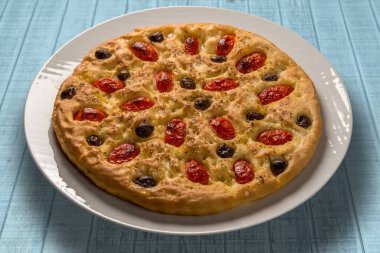 Apulian Focaccia, üstünde vişneli domates ve siyah zeytinli tipik İtalyan Bari pizzası. Açık mavi ahşap masadaki beyaz tabakta