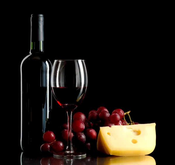 En flaske vin, en haug røde druer og et stykke ost. – stockfoto