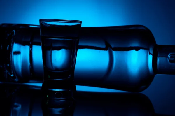 Flasche Wodka liegend mit Glas beleuchtet mit blauer Hintergrundbeleuchtung — Stockfoto