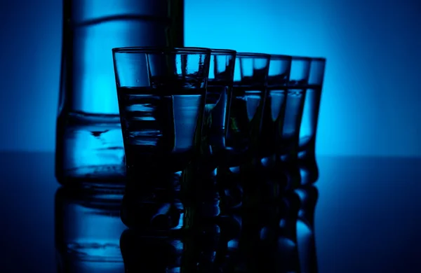 Garrafa de vodka com muitos óculos iluminados com luz de fundo azul — Fotografia de Stock