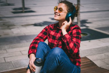 Neşeli gülümseyen kız, kulaklıkla müzik dinliyor. Şehir parkında bir bankta kahve içiyor..