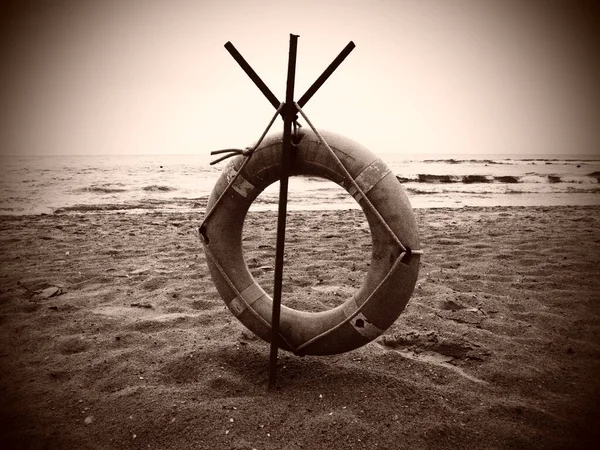 Lifebuoy on a sandy beach. Orange circle on a pole to rescue people drowning in the sea. Rescue point on the shore. Sky and sea in the background. Sepia monochrome. Dark vignetting around the edges — Stock Photo, Image