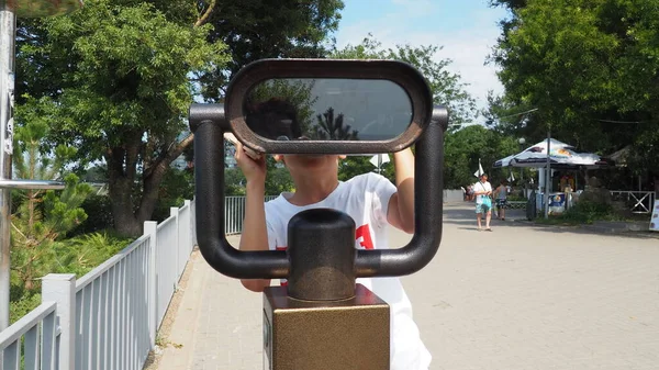 Anapa, Rusia, 23 de agosto de 2021 Un niño mira a través de un telescopio exterior. Un niño caucásico de 9 años sostiene un gran telescopio marino con sus manos. La calle de la ciudad turística y los turistas a pie — Foto de Stock