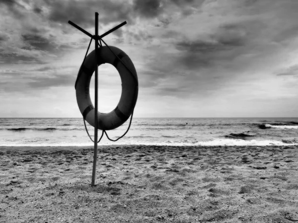 Záchranná bóje na písečné pláži. Oranžový kruh na tyči, který zachrání lidi topící se v moři. Záchranný bod na břehu. Obloha s mraky a moře v pozadí. Černobílá fotografie s vysokým kontrastem. — Stock fotografie