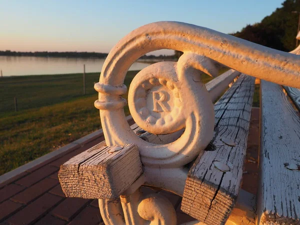 Palic, Serbia September 10, 2021. Metal bench overlooking Lake Palic. The seat is white. R letter on metal armrest. Golden hour in a quiet family resort. Tourist destination near the Hungarian border.