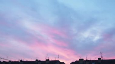 Akşam gökyüzü ve pembe bulutlar. Şehir binalarının üzerinde gün batımı. Zaman atlamalı bir atmosferde hızlandırılmış hareket. Güzel bulutlar. Gökyüzünde hızlı hareket eden bulutlar. Yavaş yavaş kararma