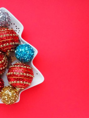 Noel ağacı şeklinde beyaz bir tabakta çok renkli Noel topları. Ağacın yarısı solda. Uzayı sağa kopyala. Kırmızı arka plan. Güzel çizgili yeni yıl dekoru ve ışıltısı.