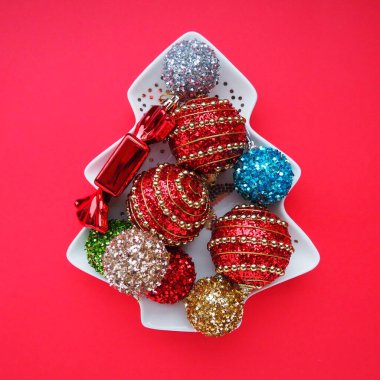 Noel ağacı şeklinde beyaz bir tabakta çok renkli Noel topları. Noel ağacı. Kırmızı arka plan ya da kırmızı renk. Güzel çizgili Noel dekoru ve ışıltılar. Düz yatıyordu. Durgun hayat