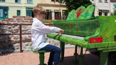 Beyaz gömlekli bir çocuk sokakta piyano çalar. 9 yaşında, beyaz, sarı saçlı bir çocuk. Piyano yeşile boyanmış. Bir parça müziğin performansı. Güneşli bir yaz gününde sokak konseri. Sırbistan.