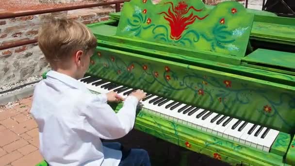 白いシャツを着た少年が通りでピアノを演奏する 9歳の白人で金髪 ピアノは緑を描いた 音楽作品の演奏 夏の晴れた日にストリートコンサート セルビア — ストック動画