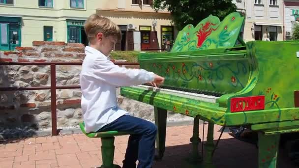 白いシャツを着た少年が通りでピアノを演奏する 9歳の白人で金髪 ピアノは緑を描いた 音楽作品の演奏 夏の晴れた日にストリートコンサート セルビア — ストック動画
