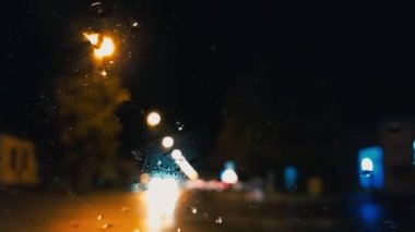 Gece araba sürüyorum. Arabanın ön camında yağmur damlaları var. Asfalt yolu. Bulanık ve odaksız etki. Camdaki damlacıklara odaklan. Gece lambaları, yaya ve bisikletçiler kavşakta.