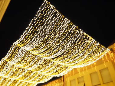 Gece gökyüzünün arka planında asılı çelenkler. Şehir sokaklarının şenlikli dekorasyonu. Sarı, beyaz ve altın renkli çelenkler ve ampuller. Yeni yıl için güzel bir geçmiş, Noel tatili