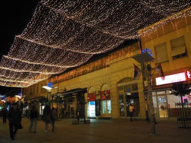 Sremska Mitrovica, Sırbistan, 24 Aralık 2021 Asılı çelenkler. Şehir sokaklarının şenlikli dekorasyonu. Sarı, beyaz ve altın renkli çelenkler ve ampuller. İnsanlar caddede dükkanların ve kafelerin önünden geçiyor..