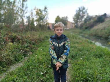 Sremska Mitrovica Sırbistan 20 Ekim 2021. 8 yaşında, yeşil ceketli yakışıklı sarışın bir çocuk sonbahar yollarında kameraya bakar. Arka planda doğa var. Bir çocuğun ciddi yüzü.