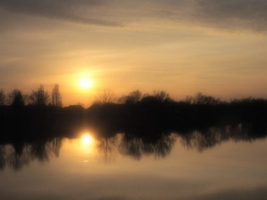 Suyun üzerinde çok güzel bir gün batımı. Nehir kıyısı ve ufuk. Parlak bulutlar akşam güneşi tarafından boyanır. Batan güneş. Sarı, turuncu, gri dramatik gökyüzü ve bulutlar. Bulanık bulanık odak.