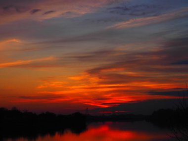 Suyun üzerinde çok güzel bir gün batımı. Nehir kıyısı ve ufuk. Parlak bulutlar, ufukta batan akşam güneşi tarafından boyanır. Kırmızı turuncu mavi dramatik gökyüzü ve bulutlar. Bulanık bulanık odak.
