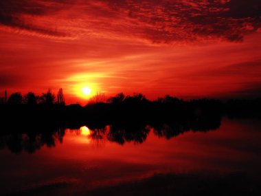 Suyun üzerinde çok güzel bir gün batımı. Nehir kıyısı ve ufuk. Parlak bulutlar, ufukta batan akşam güneşi tarafından boyanır. Kırmızı dramatik gökyüzü ve bulutlar, Güneşin sarı diski.