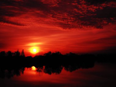 Suyun üzerinde çok güzel bir gün batımı. Nehir kıyısı ve ufuk. Parlak bulutlar, ufukta batan akşam güneşi tarafından boyanır. Kırmızı dramatik gökyüzü ve bulutlar, Güneşin sarı diski.