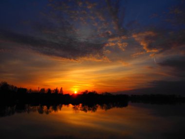 Suyun üzerinde çok güzel bir gün batımı. Nehir kıyısı ve ufuk. Parlak bulutlar, ufukta batan akşam güneşi tarafından boyanır. Kırmızı, turuncu, mavi dramatik gökyüzü bulutları. Bulanık bulanık odak.