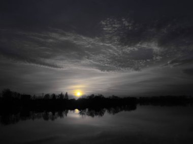 Suyun üzerinde çok güzel bir gün batımı. Nehir kıyısı ve ufuk. Güneş batıyor. Artan kontrastlı siyah beyaz bir fotoğraf. Dramatik gökyüzü ve bulut. Gecenin gelişi. Kümülüs ve sirrus bulutları