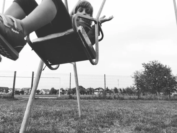 Una niña de 6-7 años balanceándose en un columpio contra el fondo de un campo de fútbol. Imagen monocromática en blanco y negro en estilo retro o vintage. Niño caucásico —  Fotos de Stock