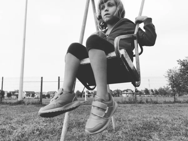 Ein 6-7 jähriges Mädchen schwingt auf einer Schaukel vor dem Hintergrund eines Fußballfeldes. Schwarz-weiß monochromes Bild im Retro- oder Vintage-Stil. Kaukasisches Kind — Stockfoto