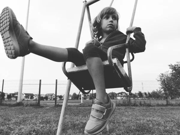 Una niña de 6-7 años balanceándose en un columpio contra el fondo de un campo de fútbol. Imagen monocromática en blanco y negro en estilo retro o vintage. Niño caucásico —  Fotos de Stock