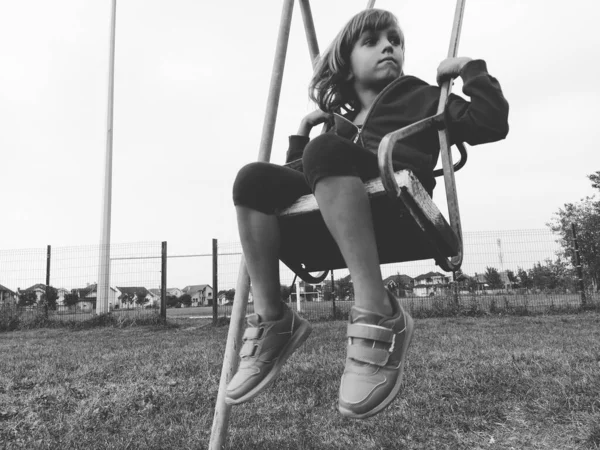 Una niña de 6-7 años balanceándose en un columpio contra el fondo de un campo de fútbol. Imagen monocromática en blanco y negro en estilo retro o vintage. Niño caucásico —  Fotos de Stock