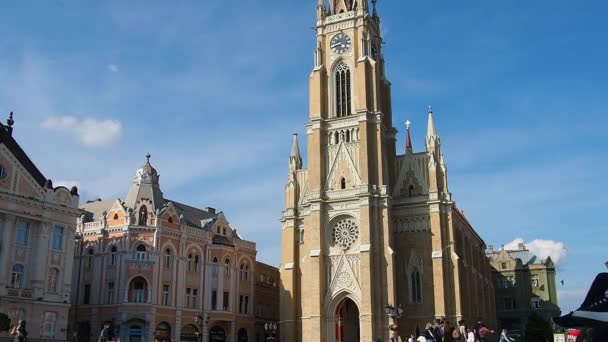 2022年4月30日，塞尔维亚诺维萨德，阳光充足的自由广场。人们，男人和女人，沿着古老的铺路石走着。圣母玛利亚教堂，哥特式。古老的旅游景点. — 图库视频影像