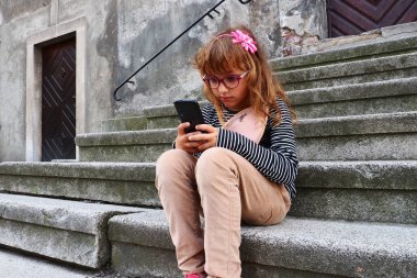 8 yaşındaki gözlüklü bir kız Petrovaradin 'deki bir kilisenin merdivenlerinde oturuyor, telefona bakıyor ve akıllı telefon kullanıyor. Şehir hayatı ve iletişim. Modern tempo. Telefon ve internet bağımlılığı.