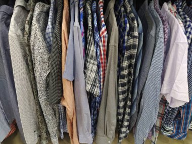Elbiseler askılarda asılı. İkinci el mağazasının çeşitli ürünleri. Askıda asılı erkek gömlekleri, müşterilere satılmaya hazır. Krizde para biriktiriyorum. Ucuz alışveriş. Damalı, çizgili, sade erkek gömlekleri..