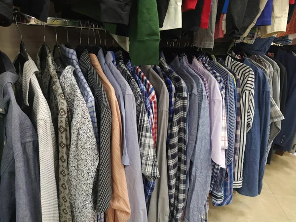 Clothes hung on hangers. Assortment of second hand store. Mens shirts hanging on a rack, ready for sale to customers. Saving money in crisis. Cheap shopping. Checkered, striped, plain mens shirts.