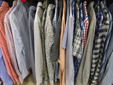 Elbiseler askılarda asılı. İkinci el mağazasının çeşitli ürünleri. Erkeklerin gömlekleri askıda asılı, müşterilere satılmaya hazır. Krizde para biriktiriyorum. Ucuz alışveriş. Damalı, çizgili, sade erkek gömlekleri..