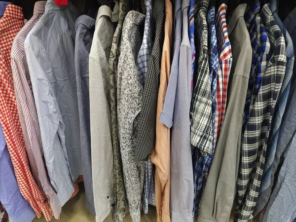 Clothes hung on hangers. Assortment of second hand store. Mens shirts hanging on a rack, ready for sale to customers. Saving money in crisis. Cheap shopping. Checkered, striped, plain mens shirts.