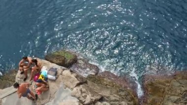 Dubrovnik, Hırvatistan, 14 Ağustos 2022 plaj elbiseli gençler, kızlar ve erkekler kıyıda güneşleniyor. Deniz dalgaları kayalara çarpıyor ve püskürtüyor. Buza plajı. Turizm, seyahat ve ekoloji.