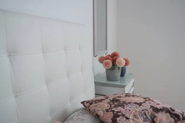 Bedroom interior design. Mirror on the wall. A bouquet of beautiful pink flowers on a white table in the bedroom. Interior Design. Bed with white upholstered headboard. Decorative pillow.