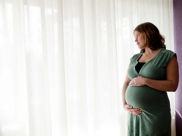 Zwangere jonge vrouw kijken uit het raam — Stockfoto