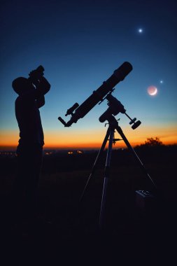 Bir adamın silueti, teleskop ve kırsal alan yıldızlı gökyüzünün altında.
