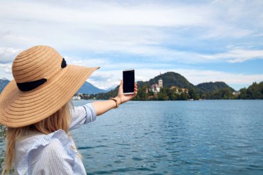 Genç, yetişkin bir turist kadın modern bir akıllı telefonla fotoğraf çekerken gölde eğleniyor..