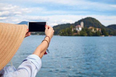 Genç, yetişkin bir turist kadın modern bir akıllı telefonla fotoğraf çekerken gölde eğleniyor..