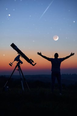 Bir adamın silueti, teleskop ve kırsal alan yıldızlı gökyüzünün altında.
