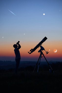 Bir adamın silueti, teleskop ve kırsal alan yıldızlı gökyüzünün altında.