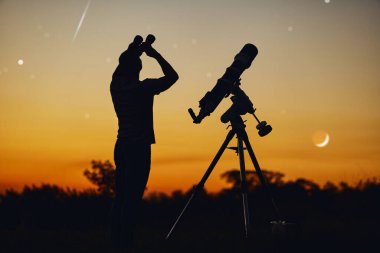 Bir adamın silueti, teleskop ve kırsal alan yıldızlı gökyüzünün altında.