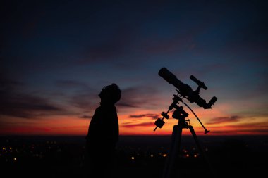 Bir adamın silueti, teleskop ve kırsal alan yıldızlı gökyüzünün altında.