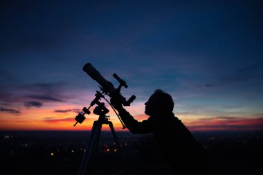Bir adamın silueti, teleskop ve kırsal alan yıldızlı gökyüzünün altında.