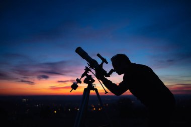 Bir adamın silueti, teleskop ve kırsal alan yıldızlı gökyüzünün altında.