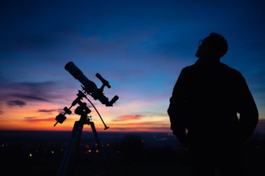 Bir adamın silueti, teleskop ve kırsal alan yıldızlı gökyüzünün altında.