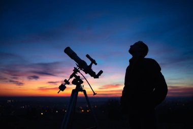 Bir adamın silueti, teleskop ve kırsal alan yıldızlı gökyüzünün altında.